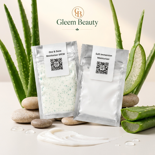Two silver pouches of Gleem Beauty moisturizers, one labeled 'One & Done Moisturizer SPF30' with green-blue speckled cream and the other 'Soft Immersion Moisturizer' with white cream, styled with aloe vera leaves, sliced aloe, smooth stones, and a dollop of cream on a light beige background.